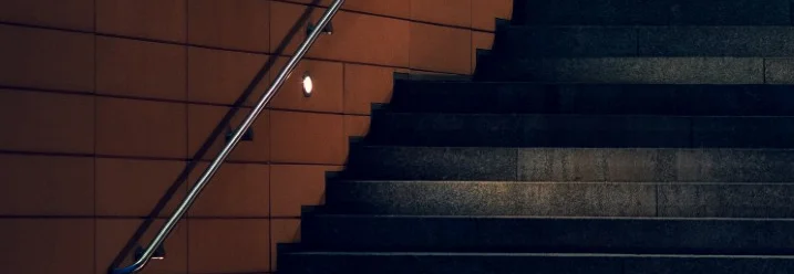 Dunkle Steintreppe mit Handlauf an einer beleuchteten, orangefarbenen Wand.